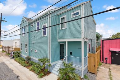 Turquoise two-story home with white trim, covered front porch and steps, tropical palm landscaping along the sidewalk, power lines overhead, and a bright pink neighboring building.
