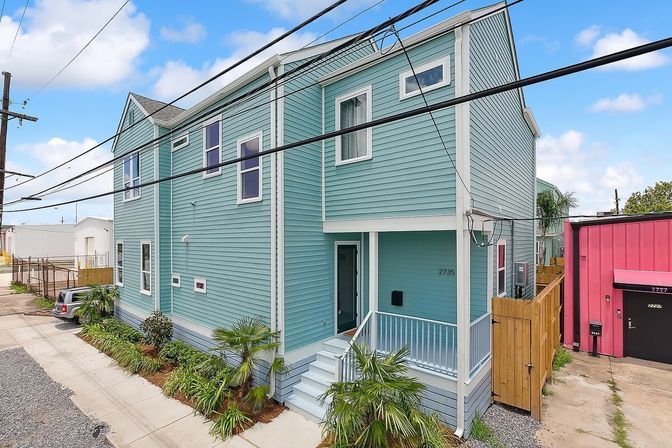 Turquoise two-story home with white trim, covered front porch and steps, tropical palm landscaping along the sidewalk, power lines overhead, and a bright pink neighboring building.