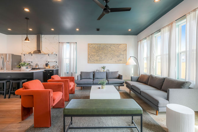Bright modern open-concept living room and kitchen with teal ceiling, two orange velvet armchairs, gray sofas, marble backsplash, large windows, wood floors, and framed map artwork.