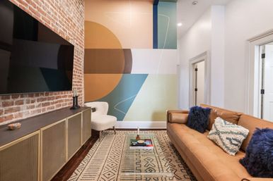 Contemporary urban living room with exposed brick and wall-mounted TV, colorful geometric mural, tan leather sofa with blue pillows, patterned rug and glass coffee table.
