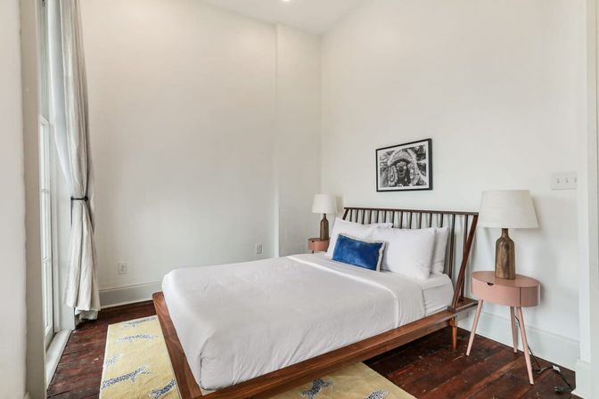 Bright minimalist bedroom with a low wooden platform bed, crisp white bedding and a blue accent pillow, pink round bedside tables with lamps, black-and-white framed art, floor-to-ceiling window with curtains, dark hardwood floors and a yellow patterned rug.