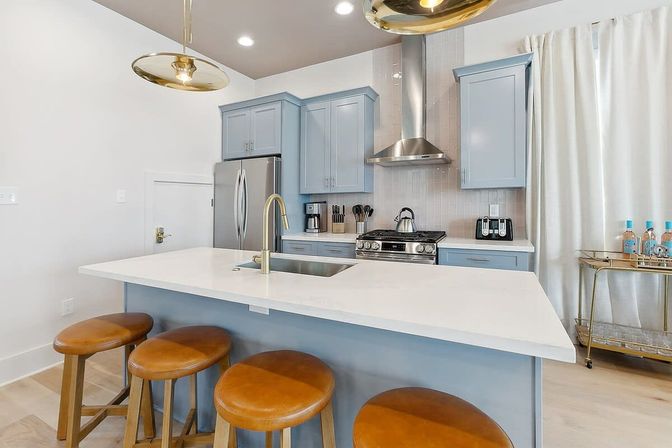 Bright modern kitchen with light blue cabinets, white quartz island and brass faucet, stainless steel refrigerator and range, four round leather bar stools, gold pendant lights and a bar cart.