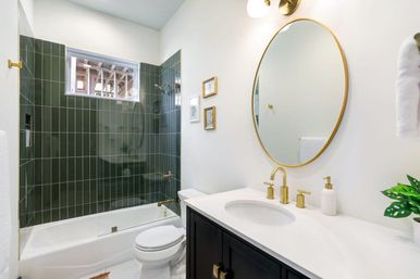 Modern bathroom with dark green vertical subway-tile shower around a white tub, brass fixtures, round gold mirror, white countertop and dark vanity
