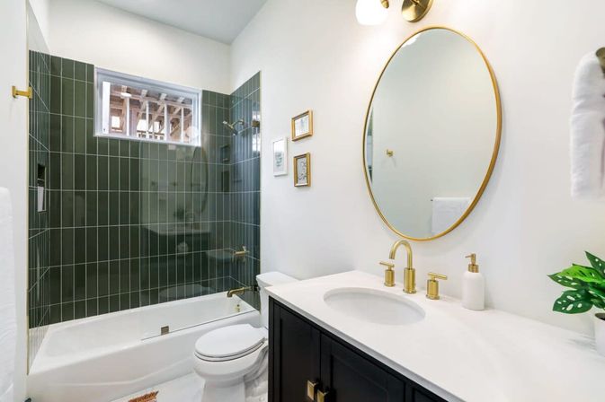 Modern bathroom with dark green vertical subway-tile shower around a white tub, brass fixtures, round gold mirror, white countertop and dark vanity