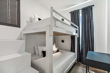 Compact modern city bedroom with white bunk bed (full lower, twin upper) with stepped access, crisp white bedding, black accent pillows, dark floor-length curtains and a small black desk.