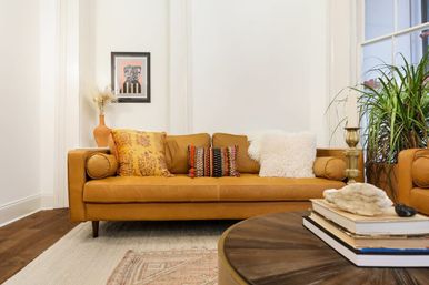 Bright living room with tan leather sofa dressed in colorful patterned and fluffy pillows, wooden coffee table with stacked books and shell, brass candleholder, potted plant by a window and framed art on white walls.
