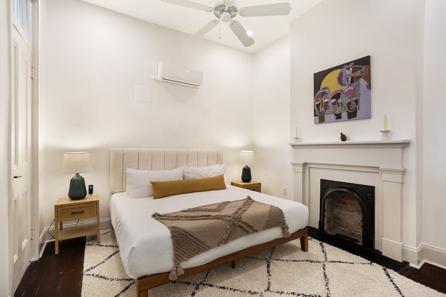 Bright cozy modern apartment bedroom with upholstered bed, wooden nightstands and lamps, ceiling fan, wall AC, patterned rug and decorative fireplace with abstract artwork.