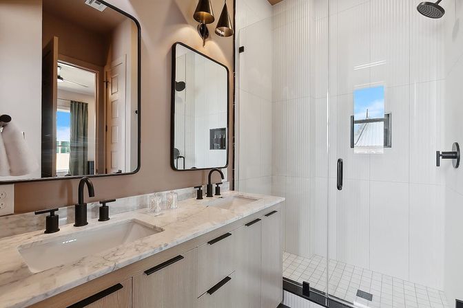 Sleek modern bathroom with double marble vanity, matte-black faucets, wood cabinets, large mirrors and glass walk-in shower with white vertical tile