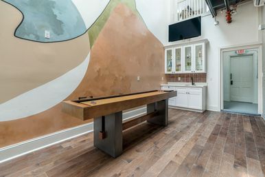 Modern indoor game room with a wooden shuffleboard table on wide-plank hardwood floors, abstract painted mural on the wall, white kitchenette with glass-front cabinets and sink, wall-mounted TV and an exit door.