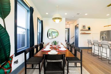 Modern open-plan dining room with long wooden table set for eight, black woven chairs, red placemats and white dinnerware, a pendant light, large windows with dark curtains, and adjacent white kitchen island on light hardwood floors.