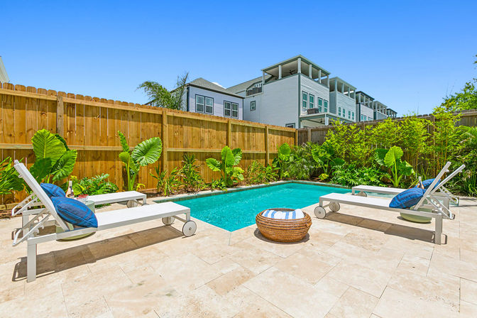 Sunny backyard swimming pool with rectangular turquoise water, four white lounge chairs with blue cushions on a travertine patio, tropical plants and wooden privacy fence with modern townhomes beyond.