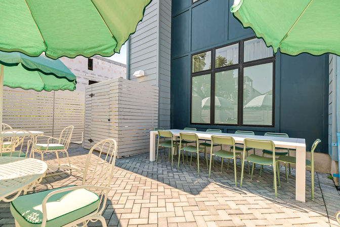 Sunlit urban outdoor patio with a long white communal table lined with green metal chairs, round bistro tables with green-cushioned seats, scalloped green umbrellas, and herringbone paver flooring beside a modern blue building.
