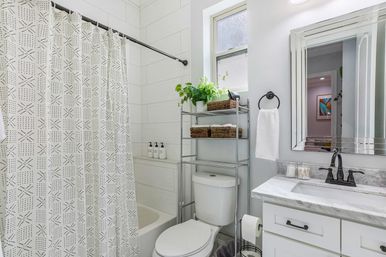 Bright modern home bathroom with patterned shower curtain, white tub and large tile, over-toilet metal shelving with green plant and wicker baskets, marble vanity with black faucet, mirror and white hand towel