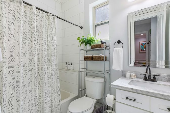 Bright modern home bathroom with patterned shower curtain, white tub and large tile, over-toilet metal shelving with green plant and wicker baskets, marble vanity with black faucet, mirror and white hand towel