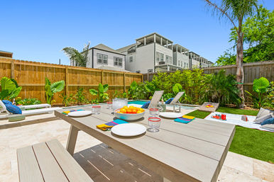 Sunny backyard patio with a wooden dining table set with plates and a bowl of oranges, lounge chairs beside a small pool, tropical plants and a wooden fence with modern townhouses in the background.
