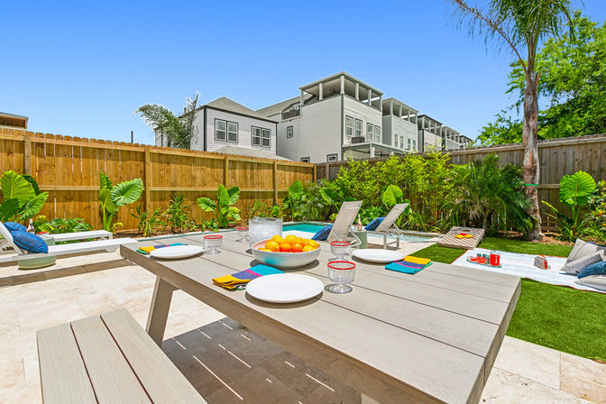 Sunny backyard patio with a wooden dining table set with plates and a bowl of oranges, lounge chairs beside a small pool, tropical plants and a wooden fence with modern townhouses in the background.