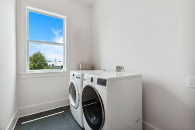 Bright, modern laundry room with white front-load washer and dryer, small utility sink, gray tile floor and a sunny window showing tree-lined views.