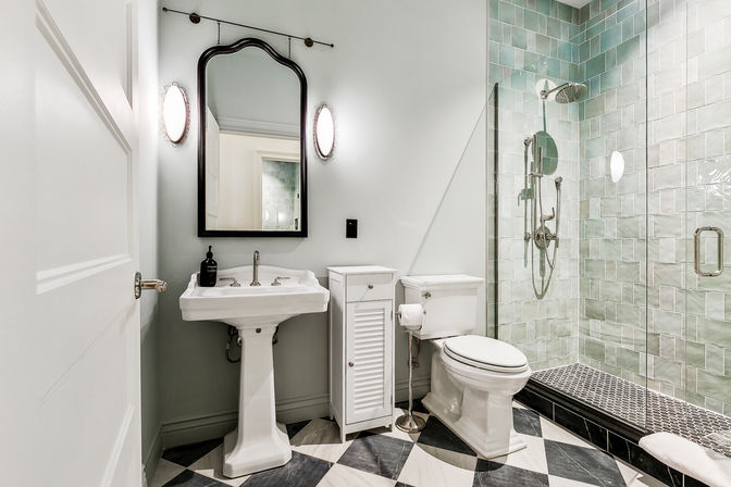 Bright, stylish modern bathroom with a pedestal sink, black-framed mirror, white toilet and slim storage cabinet, glass-enclosed shower with sea-glass green tiles and rain showerhead, and black-and-white checkered floor.