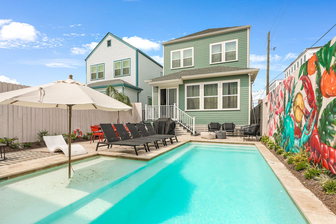 Bright backyard with an inviting aqua swimming pool, umbrella and black lounge chairs beside a two-story seafoam-green house and vibrant mural on the fence under a clear blue sky