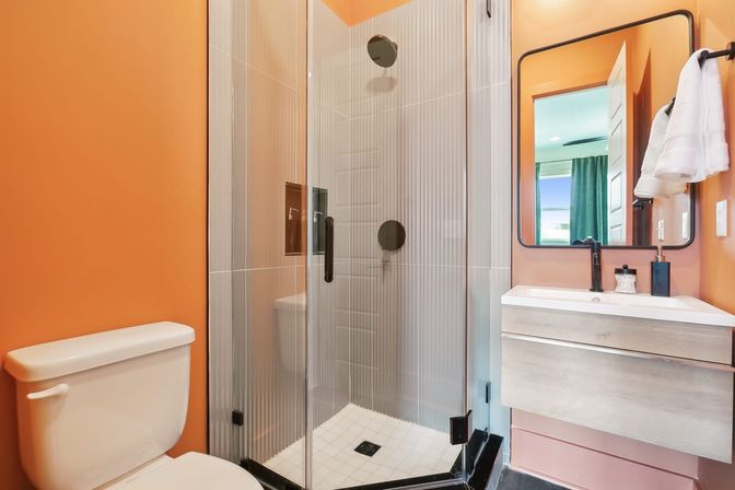 Bright modern bathroom with orange walls, glass corner shower with black fixtures, white toilet, floating wood vanity and rectangular mirror