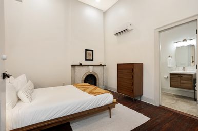 Minimalist bedroom with high ceilings and white walls, mid-century wooden bed and dresser, a vintage fireplace, hardwood floors and an en-suite bathroom