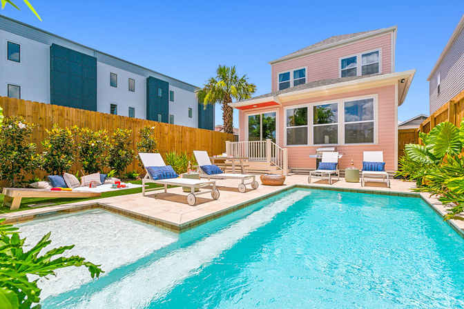 Sunny backyard pool at a pastel pink two-story house with turquoise water, chaise lounges, picnic blanket, palm tree, and wooden privacy fence