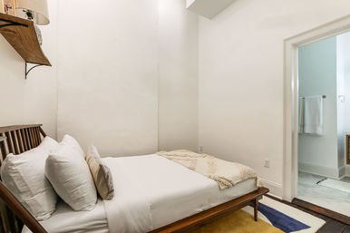 Cozy minimalist bedroom with a wooden mid-century bed frame, crisp white linens and pillows, a textured beige throw, a wall shelf lamp, colorful rug, and an open doorway to a bright tiled bathroom.
