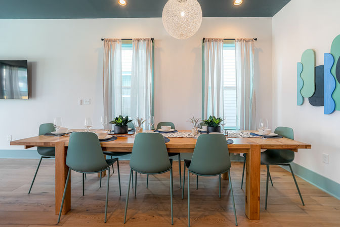 Bright modern dining room with a long wooden table set for six, teal mid-century chairs, a spherical pendant light, potted centerpieces, sheer curtains over twin windows, wood floors and abstract teal wall art.