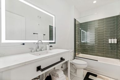 Sleek modern bathroom with white vanity and LED backlit mirror, chrome faucet, glass-enclosed bathtub-shower with green subway tile, toilet, folded towels and black hardware