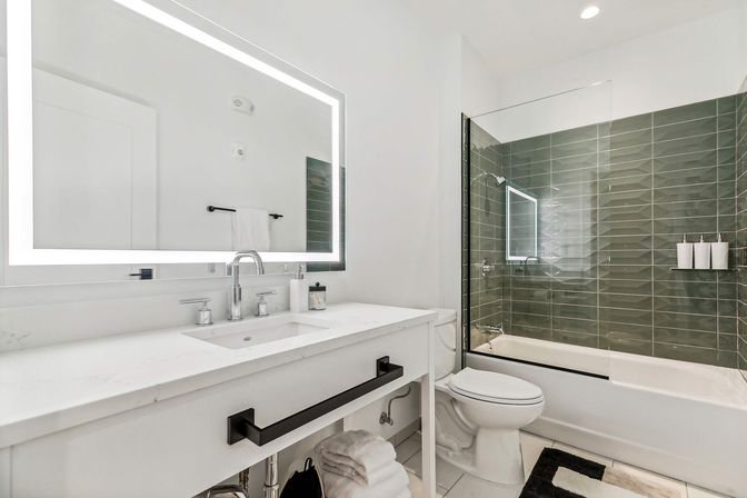 Sleek modern bathroom with white vanity and LED backlit mirror, chrome faucet, glass-enclosed bathtub-shower with green subway tile, toilet, folded towels and black hardware