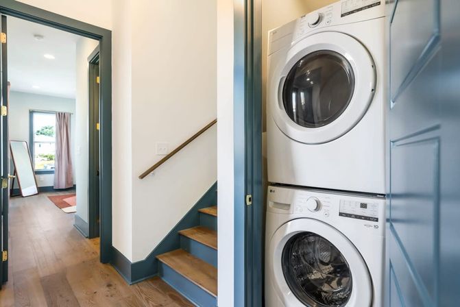 Stacked white washer and dryer tucked into a blue-trimmed closet off a bright hallway with hardwood floors, nearby wooden staircase, and a sunlit room beyond — compact laundry nook in a modern home.