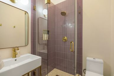 Chic compact bathroom with mauve vertical tiles in a glass walk-in shower, brass showerhead and handle, built-in shampoo niche, white rectangular sink with brass faucet, round wall sconce and white toilet.