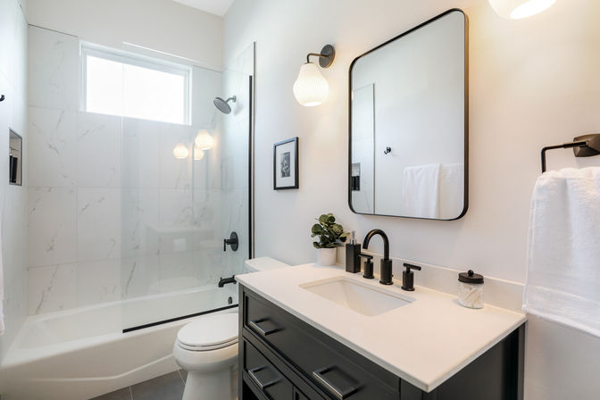 Bright modern bathroom with marble-look tiled bathtub-shower and glass door, black matte fixtures, dark vanity with white countertop and rectangular sink, black-framed mirror, wall sconce, small potted plant and neatly folded towel.