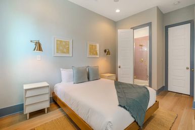 Bright modern bedroom with wooden platform bed, white linens, gray pillows and teal throw, brass wall sconces above white nightstands, hardwood floors and open door revealing pink-tiled en-suite shower