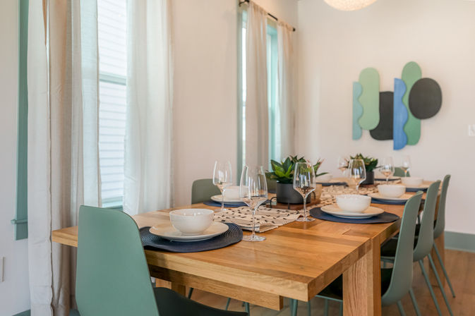 Light-filled modern dining room with a wooden table set for six, sage-green chairs, navy placemats, white bowls and wine glasses, potted succulents centerpiece, sheer curtains and abstract wall art.