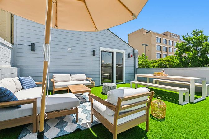 Urban rooftop patio with bright artificial grass, wooden lounge seating and cushions under a large umbrella, low coffee table, bench-style dining table, and city buildings visible in the background on a sunny day