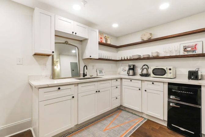 Cozy modern white kitchenette with L-shaped shaker cabinets, dark wood floating shelves, black faucet and compact appliances on marble-look countertops, framed mirror and geometric rug.