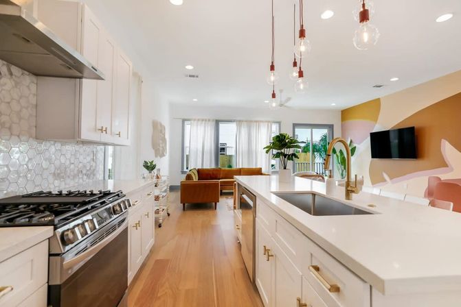 Bright modern open-plan kitchen and living area with white quartz island, brass faucet, hanging Edison bulbs, hex tile backsplash, stainless gas range, wood floors, mustard sofa and colorful geometric accent wall.