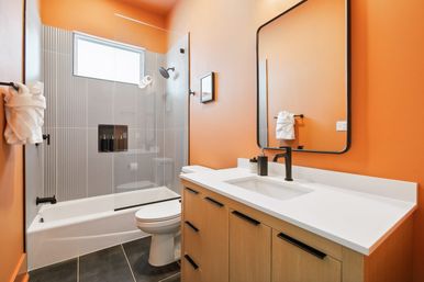 Bright orange modern bathroom with glass-enclosed tub/shower, fluted gray tile niche, matte-black fixtures, wood vanity with white countertop and black-framed mirror