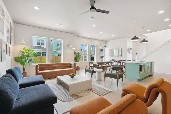 Bright modern open-plan living room and kitchen in a suburban home, featuring a teal island with pendant lights, navy and tan sofas, dining table, indoor plants, large windows, ceiling fan, and light wood floors.