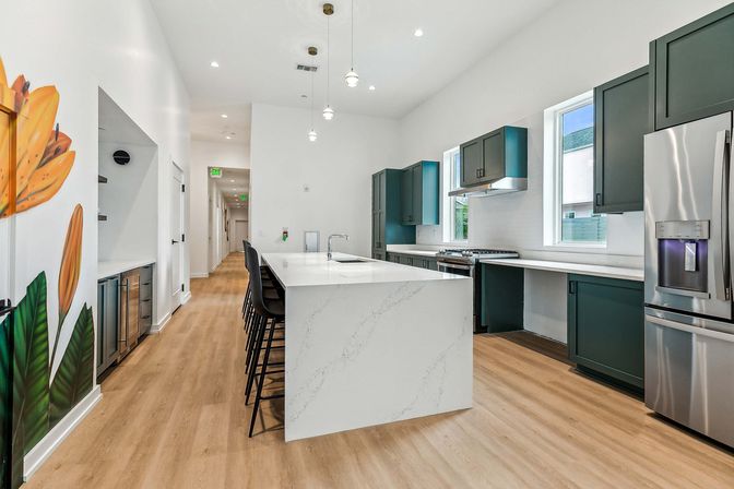 Contemporary open-concept kitchen with large white quartz waterfall island, black bar stools, dark green cabinets, stainless steel appliances, pendant lights, light wood floors and a tropical flower mural.