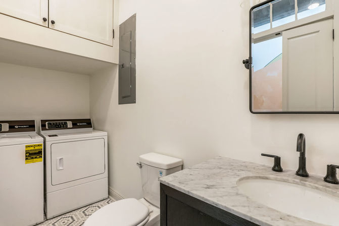 Compact laundry-bathroom with side-by-side washer and dryer, white toilet, marble vanity with black faucet, wall mirror, white cabinets and patterned tile floor.