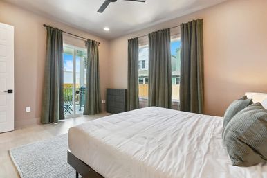 Bright modern master bedroom with white bedding and green accent pillows, floor-to-ceiling olive curtains, sliding glass door to a small balcony with chair and grill, black dresser, ceiling fan, and light hardwood floors.