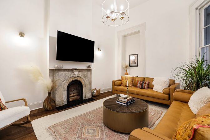 Cozy high-ceiling urban apartment living room with tan leather sofas, marble fireplace, wall-mounted TV, round coffee table, boho rug and indoor plants.