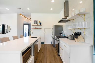 Bright modern open-concept kitchen with long white island and brass faucet, stainless steel fridge and gas range, open shelves with glassware, knife block, toaster, microwave and warm wood floors.