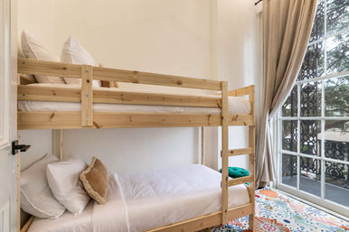 Bright cozy bedroom featuring a natural wood bunk bed with white linens and throw pillows, colorful patterned rug, and tall window with curtains opening onto a tree-lined balcony
