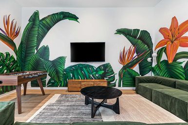Modern game lounge with oversized tropical leaf mural, green velvet sectional, foosball table, wall-mounted TV, round black coffee table and wooden media console