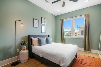 Cozy modern bedroom with mint-green walls, queen bed with white linens and gray pillows, wood floors, orange area rug, slim floor lamp and potted plant on a side table, framed wall art, ceiling fan and a sunny window with a neighborhood view.