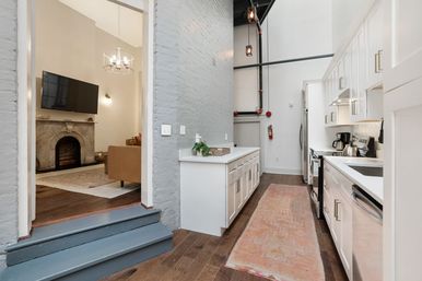 Loft-style galley kitchen with white cabinets, quartz counters and stainless appliances, long runner rug, painted exposed brick and exposed piping; opens to a living room with marble fireplace, wall-mounted TV and chandelier on hardwood floors.