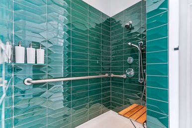 Sleek spa-like shower with glossy teal subway tiles, stainless steel grab bar, chrome handheld shower fixtures, three white wall dispensers and a folding wooden seat — modern accessible bathroom shower